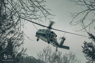 Koncepcyjna fotografia lecącego w przechyle policyjnego Black Hawka wykonana spomiędzy okalających kadr zdjęcia gałęzi drzew. W otwartych drzwiach maszyny goprowiec.