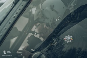 Wykonane przez szybę boczną w ujęciu profilowym zdjęcie siedzącego za sterami Black Hawka policyjnego pilota w hełmofonie na głowie. Na pierwszym planie ramię funkcjonariusza z naszywką Lotnictwa Policyjnego  KGP.
