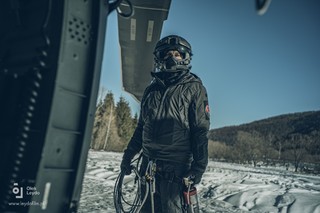 Crew Chief w umundurowaniu lotnika i hełmofonie na głowie trzyma w ręku gaśnicę stojąc przy burcie policyjnego Black Hawka. Górski zimowy krajobraz w oddali.