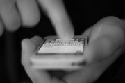 widoczna dłoń osoby trzymającej telefon komórkowy, która palcem wskazującym drugiej ręki dotyka ekranu telefonu