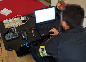 policjant Centralnego Biura Zwalczania Cyberprzestępczości siedzi przed zabezpieczonym komputerem. Obok niego, na stole, widać sprzęt elektroniczny i zabezpieczone banknoty ułożone na kupce