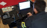 policjant Centralnego Biura Zwalczania Cyberprzestępczości siedzi przed zabezpieczonym komputerem. Obok niego, na stole, widać sprzęt elektroniczny i zabezpieczone banknoty ułożone na kupce