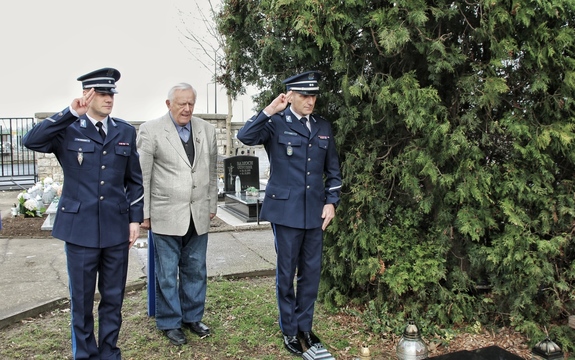 mężczyzna w ubraniu cywilnym skłania głowę i stojący obok niego dwaj funkcjonariusze salutują przed pomnikiem