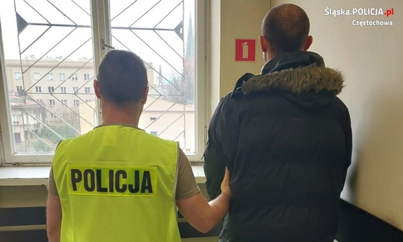Policjant z zatrzymanym stoi przed zakratowanym oknem