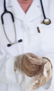Zdjęcie przedstawia fragment sylwetki osoby z personelu medycznego w białym uniformie, z założonymi lateksowymi rękawiczkami na dłoniach i słuchawkami zwisającymi z szyi.
