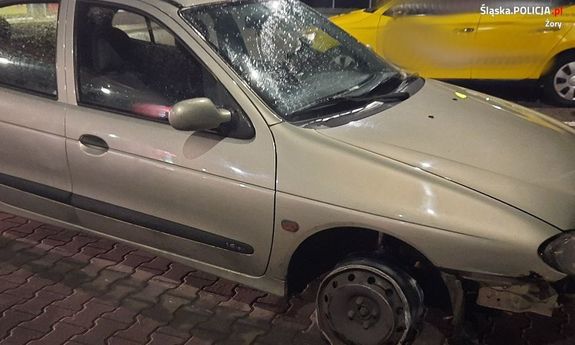 Zdjęcie przedstawia stojący bokiem pojazd  kolorze szarym, który ma stłuczoną przednią szybę i uszkodzone prawe przednie koło.