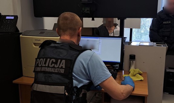 Policjant w kamizelce z napisem Policja na plecach siedzi przy komputerze - widok z tyłu