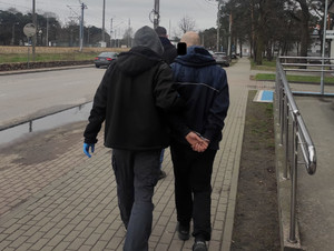 Zdjęcie przedstawi dwie osoby ubrane na ciemno idące chodnikiem. Osoba idąca po lewej stronie przytrzymuje idącego po lewej na lewe przedramię. Osoba idącą po prawej stronie ma kajdanki założone na rękach trzymanych z tyłu.