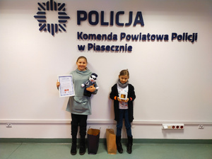 dwie dziewczynki stojące na tle napisu Komenda Powiatowa Policji w Piasecznie, w rękach trzymają nagrody