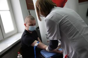 Zdjęcie przedstawia pielęgniarkę w uniformie, która pobiera krew od mężczyzny siedzącego na krześle. Oboje mają maseczki ochronne na twarzy.