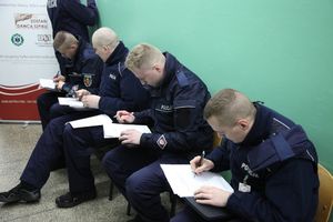 Zdjęcie przedstawia czterech umundurowanych policjantów, którzy siedzą na krzesłach w jednym rzędzie i wypełniają dokumenty.