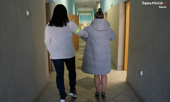 Zdjęcie przedstawia kobietę o czarnych włosach, która prowadzi po swojej prawej stronie zatrzymaną kobietę. Obie idą korytarzem, stoją tyłem do obiektywu aparatu.