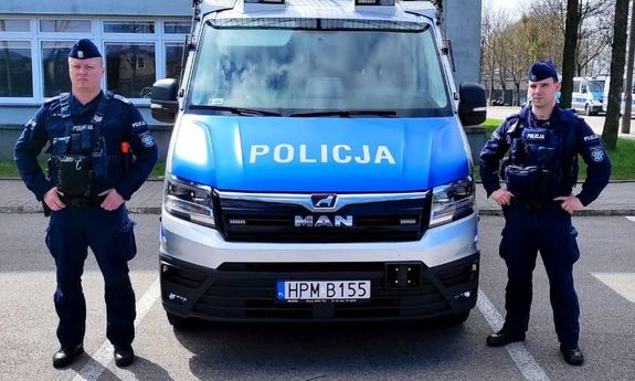 Zdjęcie przedstawia dwóch umundurowanych policjantów, którzy stoją po obu stronach oznakowanego radiowozu.