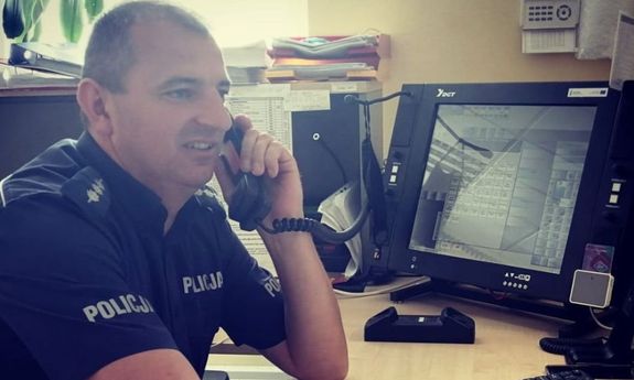 Policjant w mundurze, siedzi przy biurku na którym stoją ustawione monitory komputera, policjant trzyma przy uchu słuchawkę do telefonu