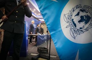 częściowo widoczny mężczyzna trzymający flagę Interpolu a za nim widoczni mężczyźni stojący przy stole prezydialnym