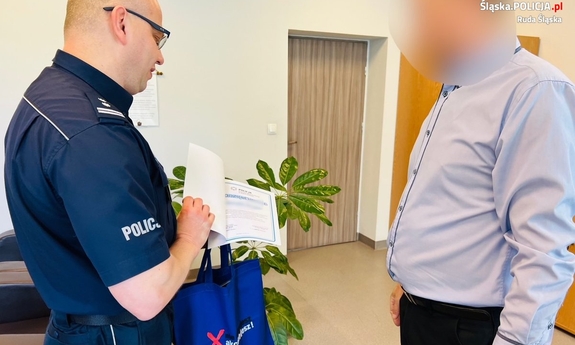 zdjęcie ze spotkania - policjant z mężczyzną w pokoju biurowym, policjant wręcza mężczyźnie dyplom i upominek w niebieskiej torbie