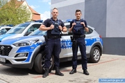 Na zdjęciu dwaj policjanci stojący przy radiowozie