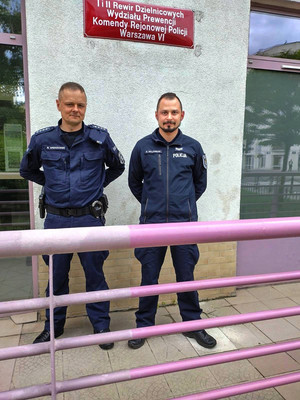 dwaj umundurowani policjanci stoją obok siebie przed budynkiem i patrzą wprost do obiektywu aparatu. Nad ich głowami, na ścianie budynku umieszczona jest czerwona tablica z białym napisem: Ii II Rewir Dzielnicowych Wydziału Prewencji Komendy Rejonowej Policji Warszawa VI