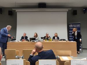 prelegenci Ogólnopolskiej Konferencji Światowej Organizacji Zdrowia siedzą za stołem prezydialnym. Jedna z nich przemawia do zebranych na sali