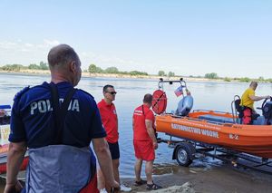 policjant oraz ratownicy wodni na brzegu rzeki, jeden z ratowników znajduje się na wodowanej łodzi