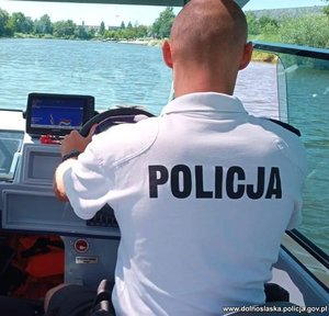 policjant w białej koszulce z napisem policja kieruje motorówką wodną, w tle widać rzekę i wybrzeże