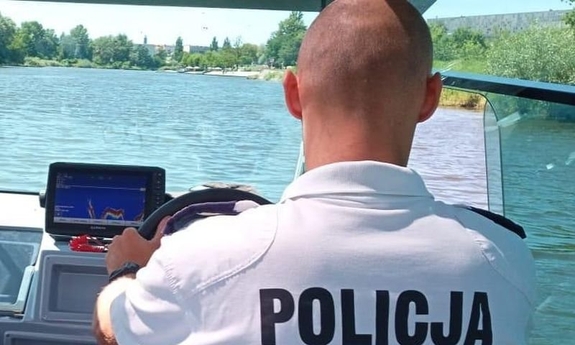 policjant w białej koszulce z napisem policja kieruje motorówką wodną, w tle widać rzekę i wybrzeże