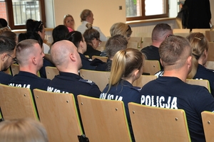 Zdjęcie pleców siedzących w rzędach krzeseł policjantów - zbliżenie na napis &quot;policja&quot;.