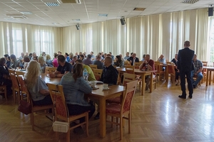 Trzy rzędy stołów z siedzącymi osobami. Przed nimi lektor.