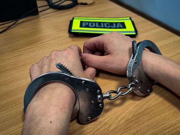 Kajdanki na rękach mężczyzny. W tle naszywka z napisem Policja