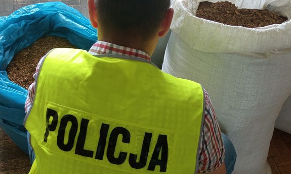 widok z tyłu na ubranego po cywilnemu policjanta w żółtej kamizelce z napisem Policja. przed funkcjonariuszem widoczne są worki wypełnione tytoniem