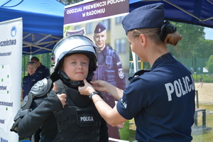 Policjantka pomaga zapiąć kask chłopcu w stroju policyjnym, w tle namioty profilaktyczne Policji.