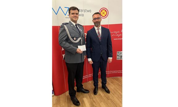 umundurowany policjant pozuje do zdjęcia trzymając legitymację honorowego dawcy krwi zasłużonego da zdrowia narodu. Obok niego stoi mężczyzna w garniturze
