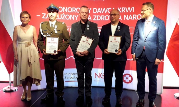 czterej mężczyźni i kobieta stoją na tle baneru w biało-czerwonych kolorach, trzej mężczyźni trzymają listy gratulacyjne oprawione w ramki