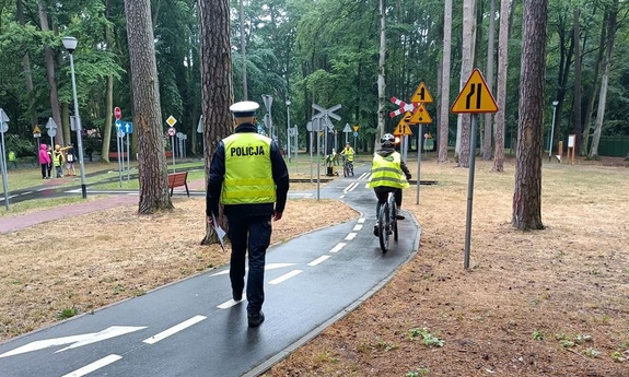policjant w żółtej kamizelce z napisem policja, w głębi dziecko w kasku na rowerze pokonujące tor przeszkód