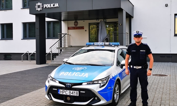 umundurowany policjant stoi przy radiowozie, w tle budynek jednostki Policji