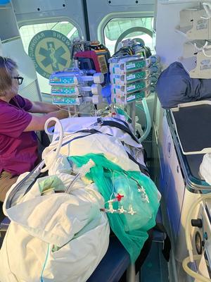 pacjent na noszach we wnętrzu karetki pogotowia, obok niego pielęgniarka, obok noszy aparatura monitorująca stan zdrowia pacjenta