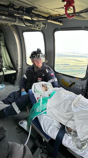 Ratownik siedzi przy pacjencie na noszach w lecącym śmigłowcu. Przez okno widać teren, nad którym leci śmigłowiec.