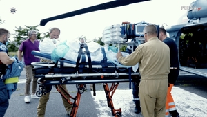 Pacjent na specjalnym łóżku na kołach obok śmigłowca. Wokół niego ratownicy i policyjni lotnicy