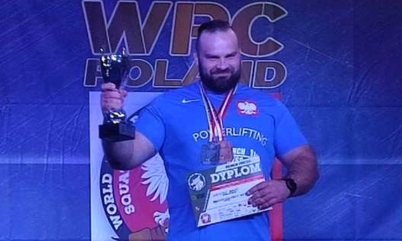 aspirant sztabowy Krzysztof Bana na podium mistrzostw w prawej ręce wzniesionej do góry trzyma puchar, w lewej ręce trzyma dyplom, na szyi ma zawieszony medal