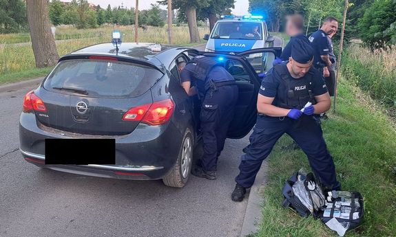 policyjny radiowóz i auto osobowe stoją na jezdni. Przy aucie widać trzech policjantów, z których jeden stoi na poboczu z ubraną po cywilnemu osobą, drugi trzyma opatrunek a przed nim na trawie leży apteczka, a trzeci nachyla się nad siedzeniem pasażera z tyłu.