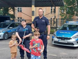 policjant i policjantka z dwójką odnalezionych dzieci stoją na parkingu policyjnym, w tle stoją dwa radiowozy policyjne