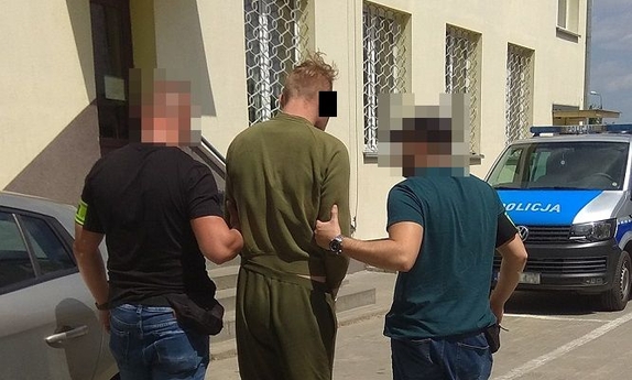dwaj policjanci prowadza zatrzymanego mężczyznę zakutego w kajdanki, w głębi radiowóz policyjny