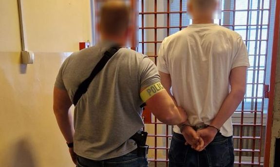 ubrany po cywilnemu policjant z opaską na ręce z napisem Policja, stoi z zatrzymanym przed wejściem do pomieszczenia dla osób zatrzymanych