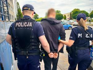 policjant i policjantka prowadzą zakutego w kajdanki mężczyznę, w tle samochody zaparkowane na parkingu