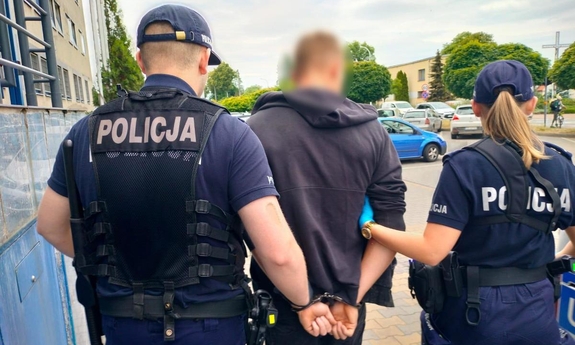 policjant i policjantka prowadzą zakutego w kajdanki mężczyznę, w tle samochody zaparkowane na parkingu