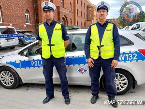 dwaj umundurowani policjanci ruchu drogowego w żółtych kamizelkach stoją przed radiowozem