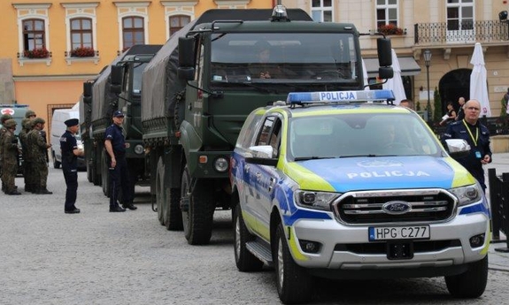 Radiowóz policyjny, a za nim stoją wojskowe samochody ciężarowe. Obok nich policjanci i żołnierze. W tle budynki
