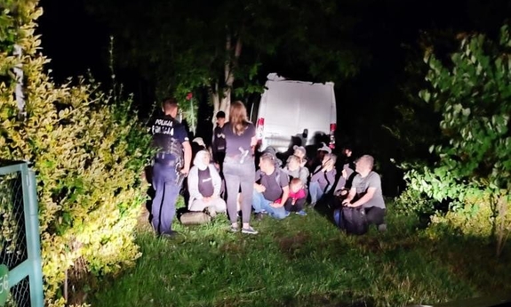 Policjanci, pogranicznicy oraz nielegalni imigranci W tle stoi biały bus. Pora nocna.