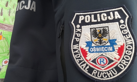 Emblemat Wydziału Ruchu Drogowego Komendy Powiatowej Policji w Oświęcimiu na mundurze, w tle fragment rysunku dziecka