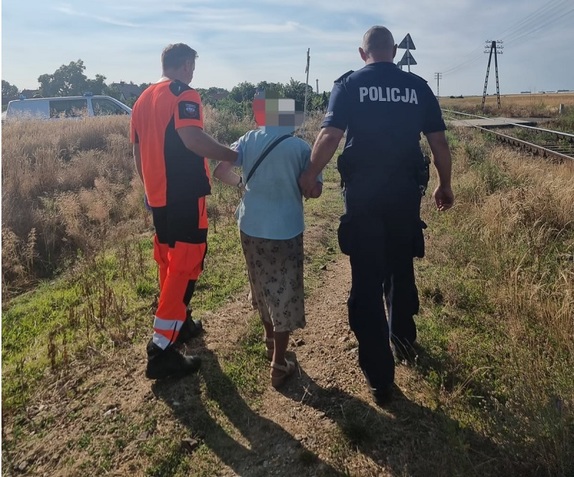 Policjant i ratownik medyczny prowadzą pod pachę starszą kobietę